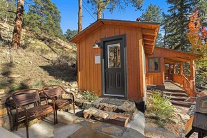 A wooden cabin with a door and steps leading up to it, surrounded by trees and a bench.