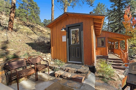 A wooden cabin with a door and steps leading up to it, surrounded by trees and a bench.