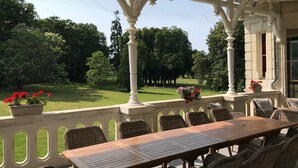 Uma área de estar coberta ao ar livre, com mesa e cadeiras, rodeada por colunas brancas e vista para um gramado e árvores bem cuidados.