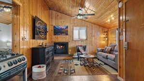 A cozy cabin interior with a fireplace, wooden walls, a sofa, and a dining area.