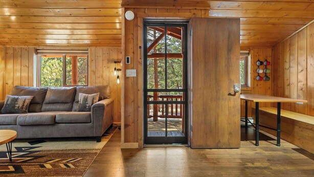 A wooden cabin interior with a sofa, a dining table, and a door leading to an outdoor deck.