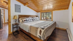 A bedroom with a wooden ceiling, a bed with a forest-themed bedspread, a nightstand with a lamp, and a window with a view of trees. - Cabin 10- King Cabin, Mins to RMNP! (Estes Park)