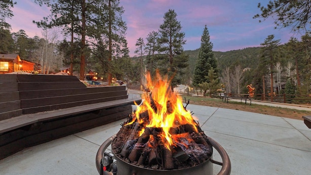 A fire pit with logs, surrounded by a concrete patio, in a wooded area with a wooden deck and a building in the background. - Cabin 10- King Cabin, Mins to RMNP! (Estes Park)