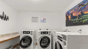 A laundry room with three washers and dryers, a guest laundry sign, a surveillance sign, and a large landscape painting on the wall. - Cabin 10- King Cabin, Mins to RMNP! (Estes Park)