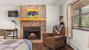 Une pièce chaleureuse avec une cheminée, un fauteuil confortable, un lit recouvert d’une courtepointe et un tableau représentant un ours accroché au mur.