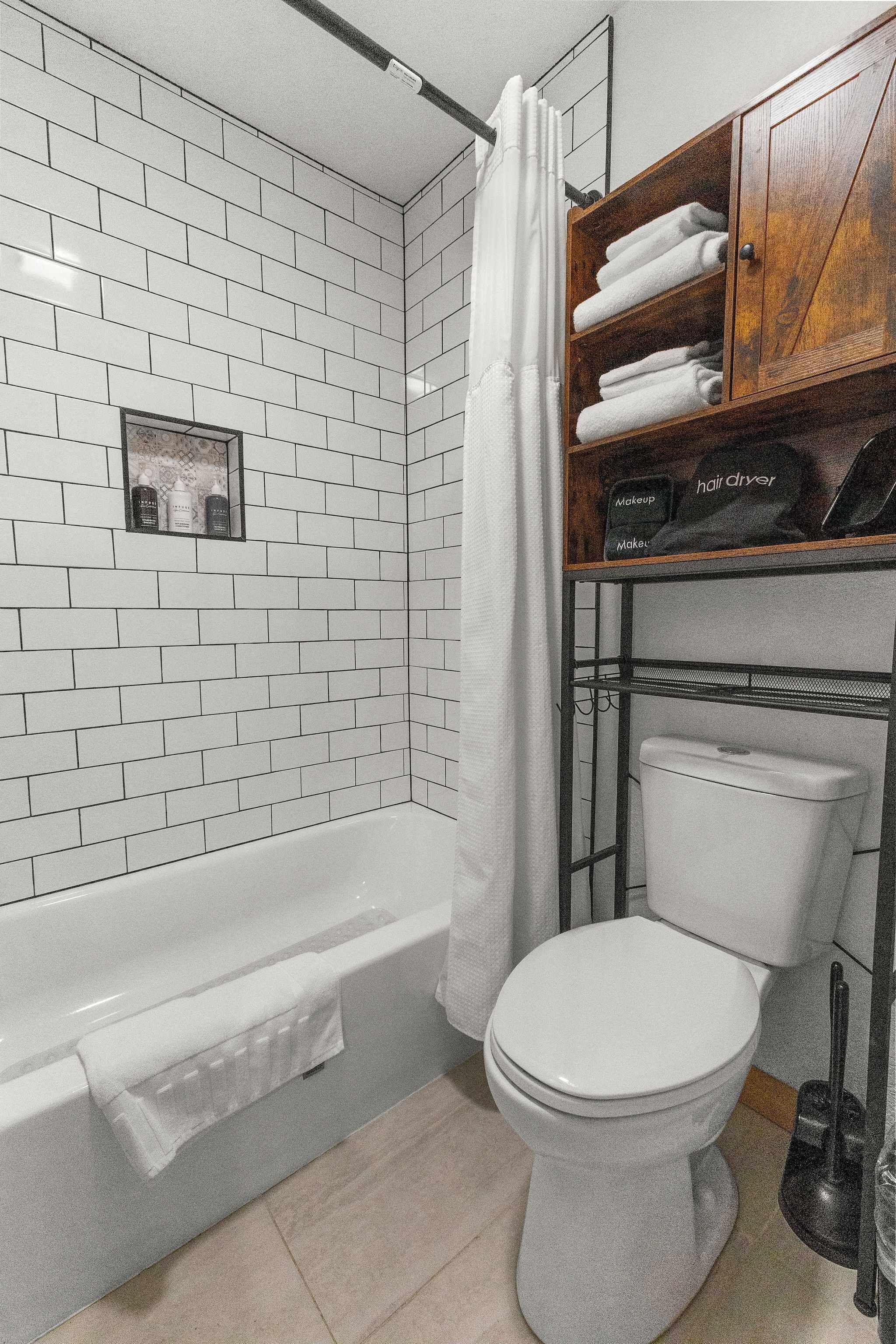 Ein Badezimmer mit weißen Fliesen, einer weißen Badewanne, einer weißen Toilette und einem Holzregal mit Handtüchern und Haarpflegeprodukten.