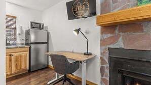 Een compacte keuken met een magnetron, koelkast en een kleine eettafel met een stoel. Achtergrond is een open haard zichtbaar, en een aan de muur bevestigde televisie toont een logo.
