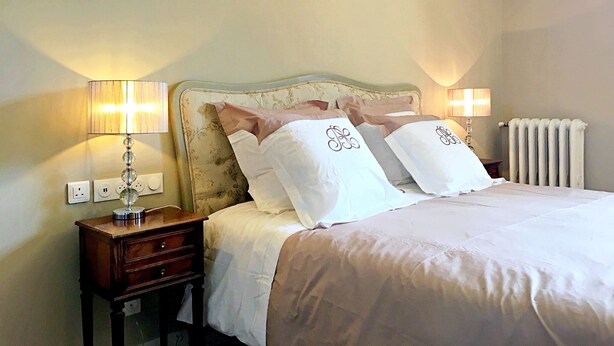 Cama bem arrumada com lençóis brancos e almofadas decorativas, mesa de cabeceira com abajur e radiador na parede.