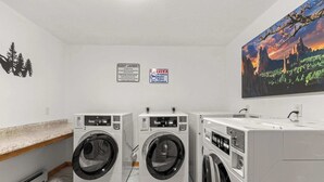 A laundry room with three washers and dryers, a guest laundry sign, a surveillance sign, and a large landscape painting on the wall. - Cabin 18 - King Bed, Mountain views! Hotel Style! (Estes Park)