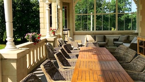 Een overdekt terras met rieten meubilair, een lange houten tafel en uitzicht op groen door grote ramen.