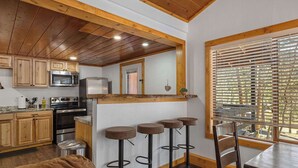 A kitchen with wooden cabinets, a microwave, a refrigerator, and a wooden ceiling. There are four stools and a table with chairs.