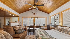 A spacious room with a high ceiling, wooden beams, a ceiling fan, a large bed with a patterned bedspread, a brown leather sofa, a dining table with chairs, and large windows with blinds.