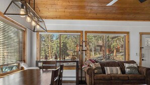 A cozy living room with a wooden dining table, a brown sofa, and large windows with blinds.