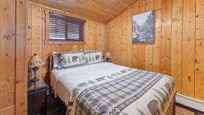 Un dormitorio de cabaña de madera con una cama, dos mesitas de noche, un ventilador de techo, una ventana con persianas y un cuadro en la pared.