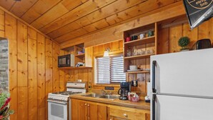 Cocina de cabaña de madera con estufa, lavabo, refrigerador y estantes con vajilla y cafetera.
