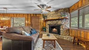 A cozy living room with a stone fireplace, wooden walls, a leather sofa, and a ceiling fan.