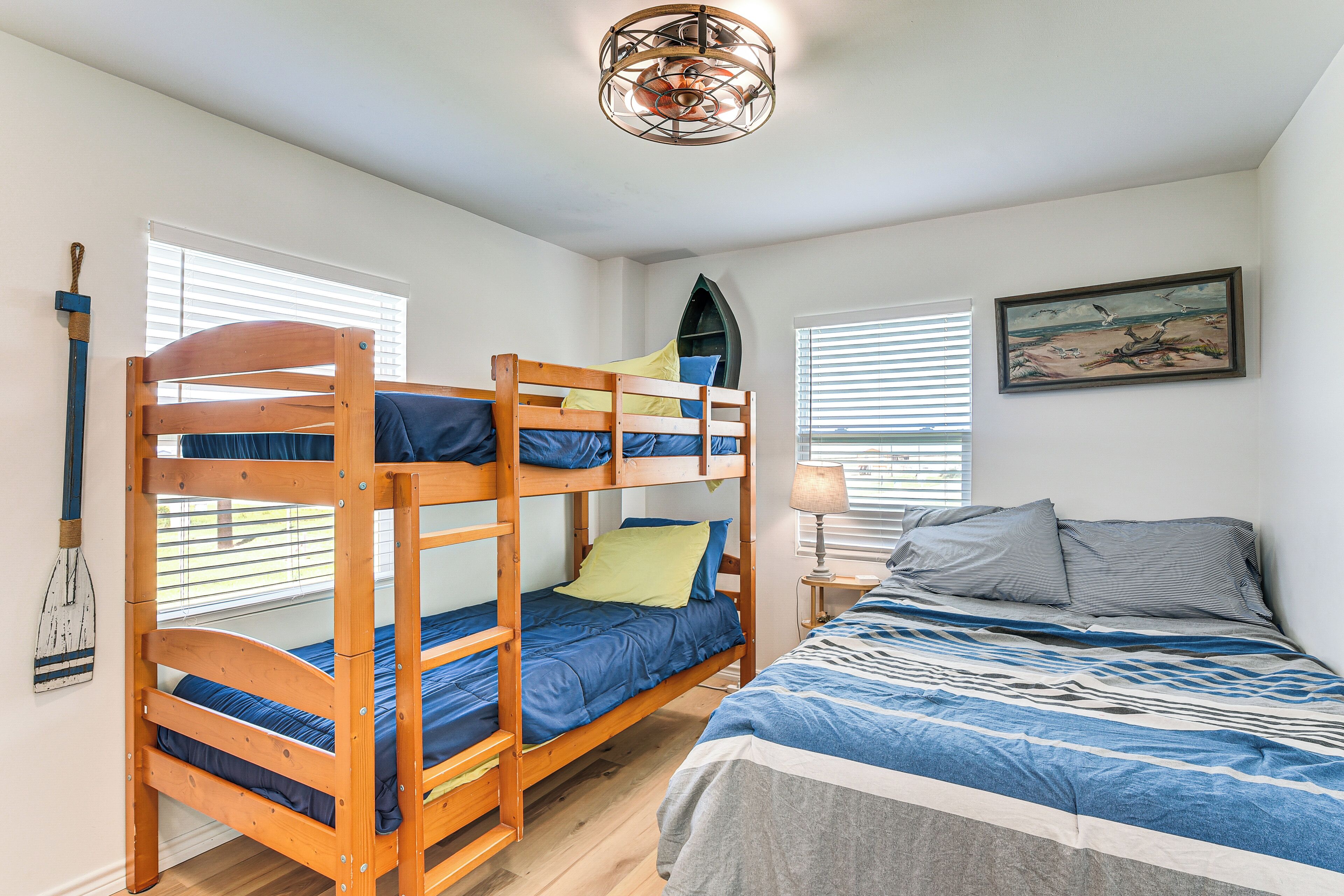 A bunk bed room with a wooden ladder, a single bed with a patterned bedspread, a wall-mounted painting, and a ceiling light fixture.