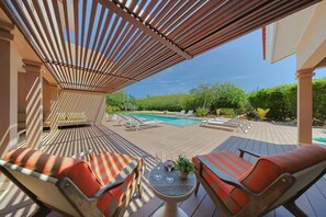 A covered outdoor area with a pool, lounge chairs, and a table with a glass of wine.