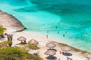 Ein Strand mit klarem türkisblauem Wasser, Menschen beim Schwimmen und Sonnenbaden sowie Reetdachschirmen.