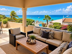 A balcony with a sofa, chairs, and a coffee table, overlooking a beach with boats and clear blue water.