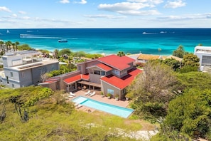 A modern two-story house with a red roof, a swimming pool, and a view of the ocean.