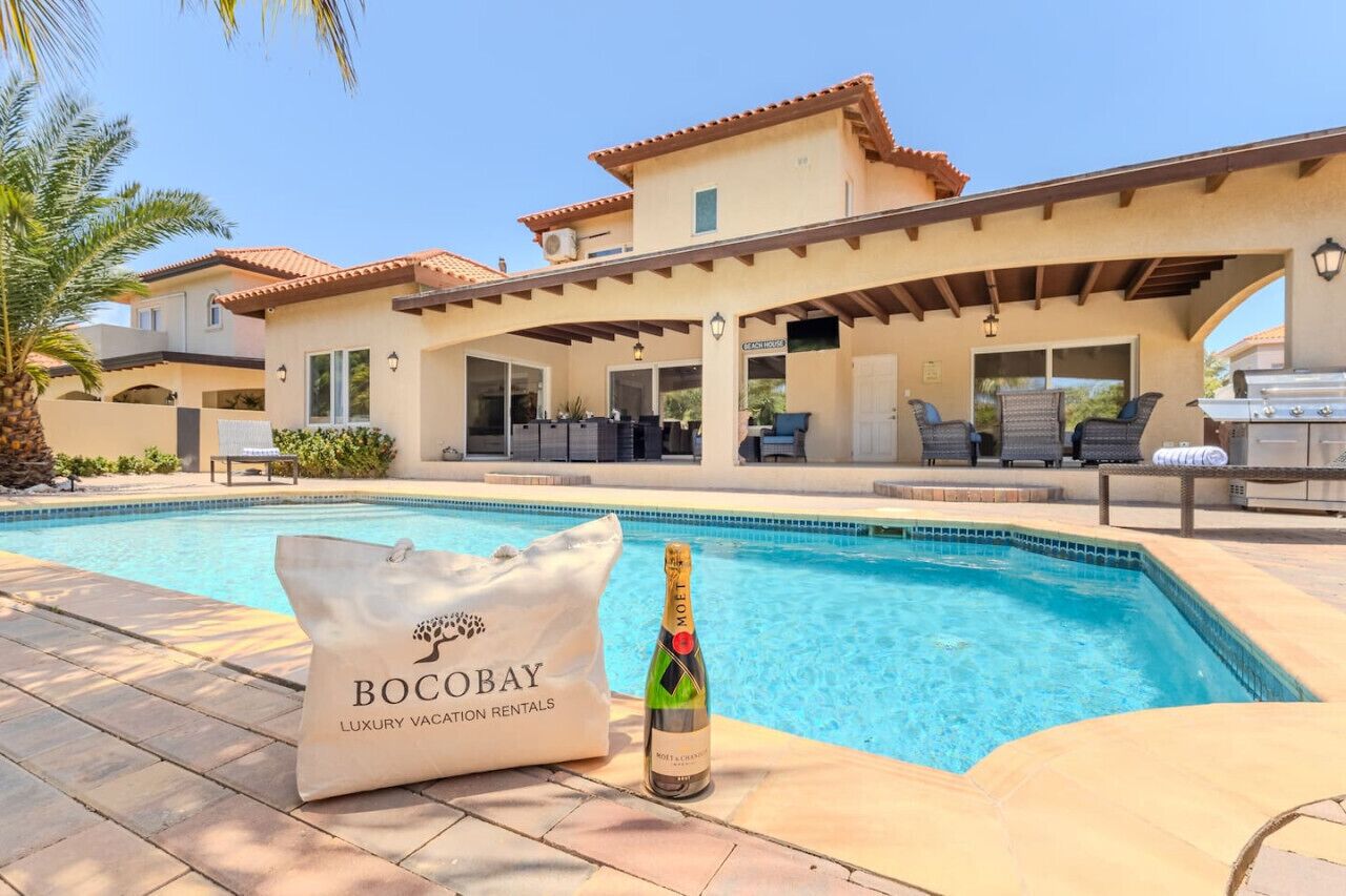 A poolside area with a bottle of champagne, a bag labeled 'BOCOBAY LUXURY VACATION RENTALS,' and a covered patio with outdoor furniture.