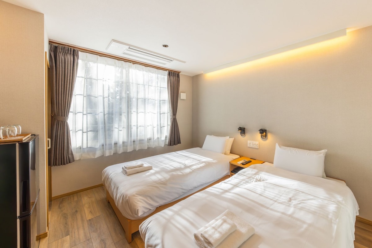 A hotel room with two beds, a bedside table with a lamp, a small desk, and a refrigerator.