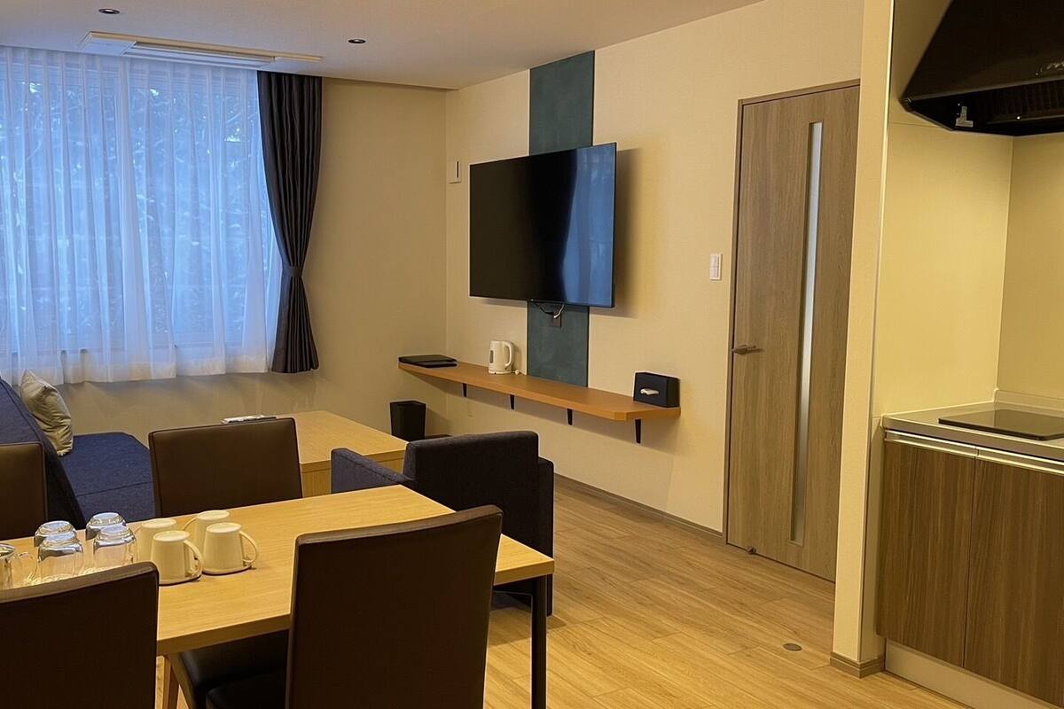 A hotel room with a dining area, a flat-screen TV mounted on the wall, a kitchen area with a refrigerator, and a window with curtains.