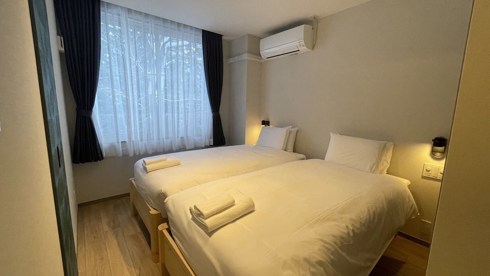 A hotel room with two beds, a window with sheer curtains, an air conditioning unit, and a bedside table with a lamp.