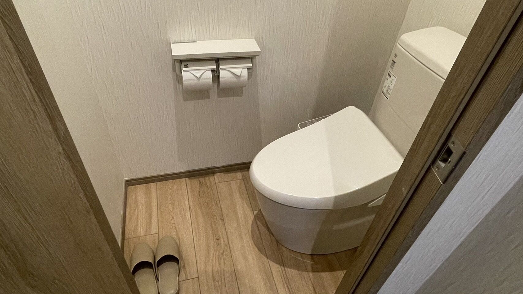 Une petite salle de bain avec des toilettes, un porte-rouleau de papier toilette et une paire de chaussons posés sur le sol.