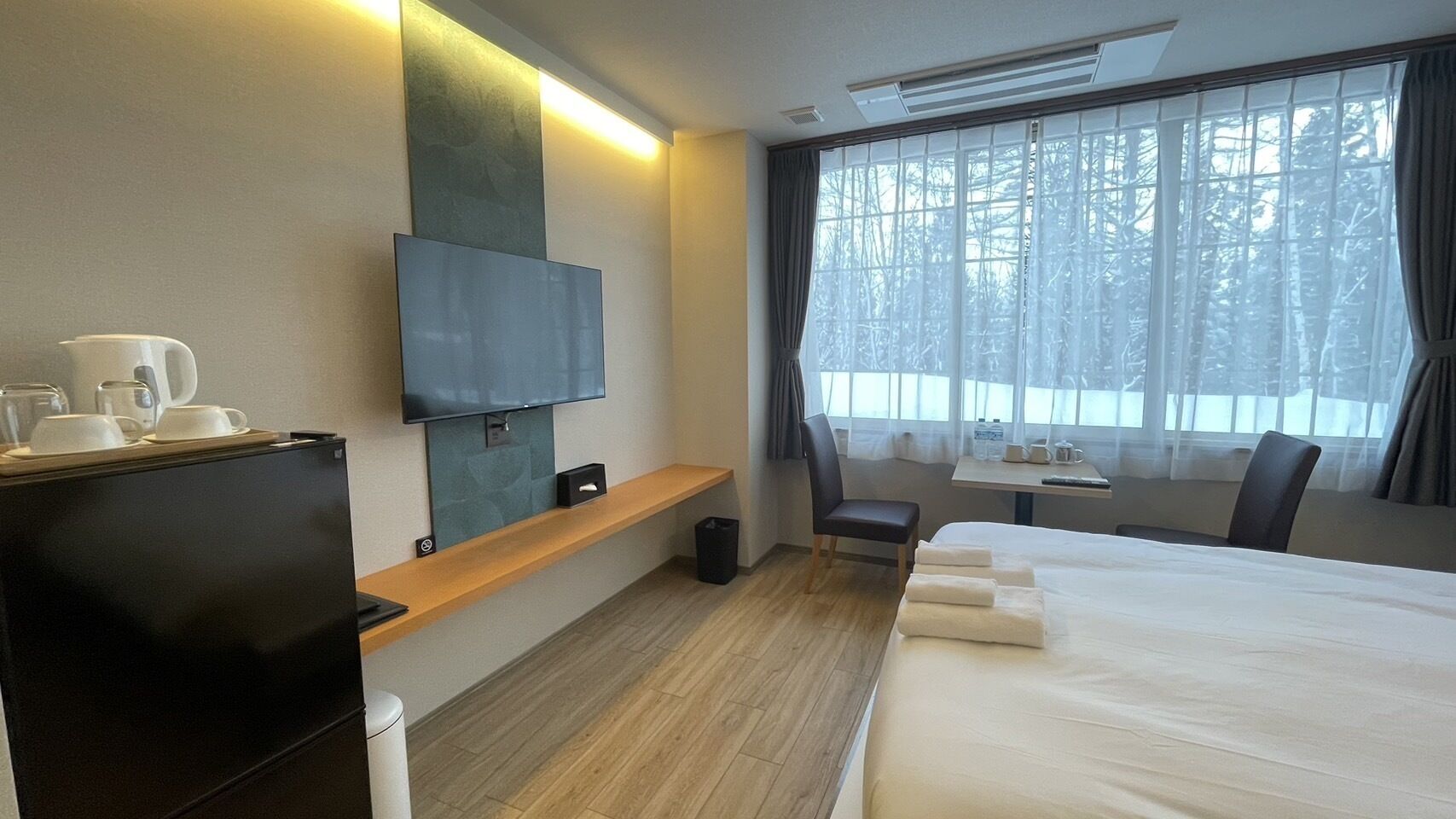 Habitación de hotel con cama, televisor montado en la pared, escritorio con silla y ventana con cortinas.