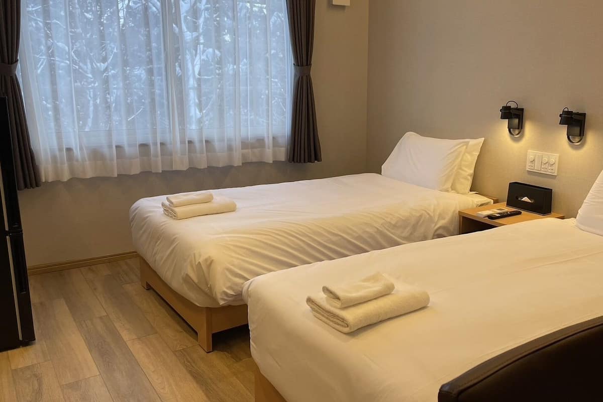 A hotel room with two beds, a wooden floor, a window with sheer curtains, and a wall-mounted light fixture.