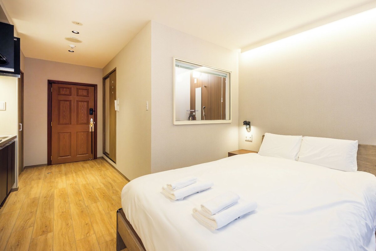 A hotel room with a bed, a wooden floor, a door, a mirror, and a nightstand with a lamp.