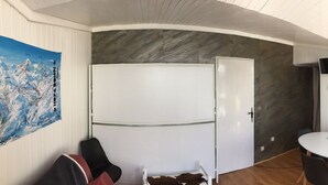 Una habitación con un refrigerador blanco, una puerta blanca, una mesa redonda con sillas, un televisor montado en la pared y un armario con puertas de vidrio.