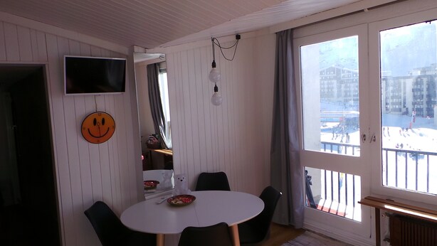 Un comedor con una mesa redonda, sillas, un reloj con cara sonriente, un televisor y una ventana que ofrece vistas a edificios y un paisaje nevado.