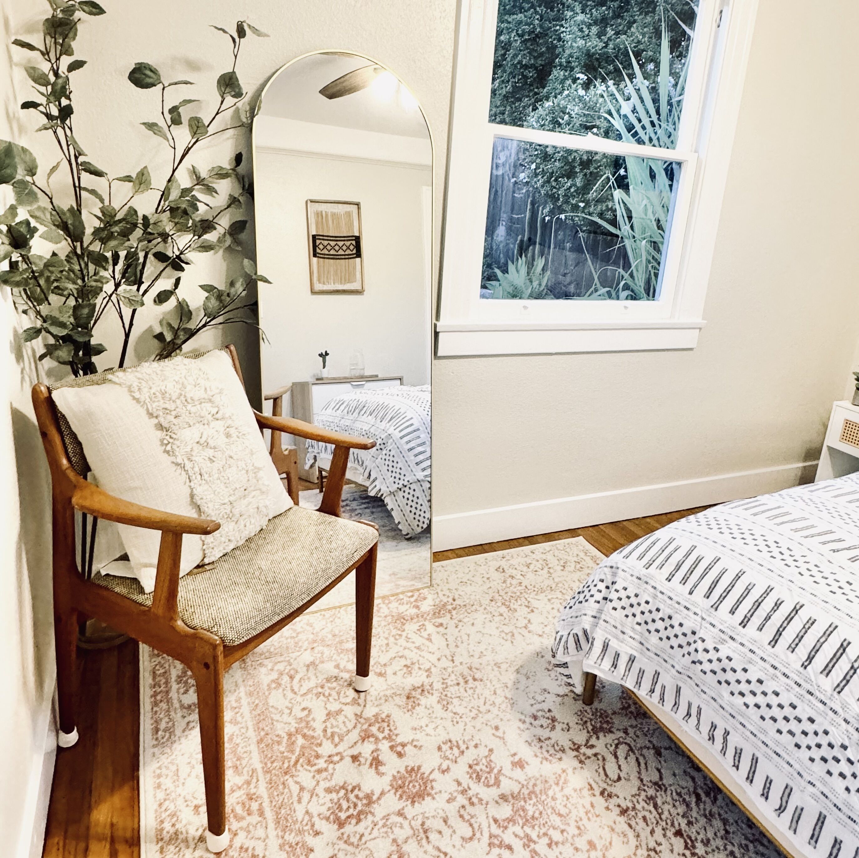A bedroom with a wooden chair, a large mirror, a bed with patterned bedding, a window with a view of greenery, and a small table with a plant.