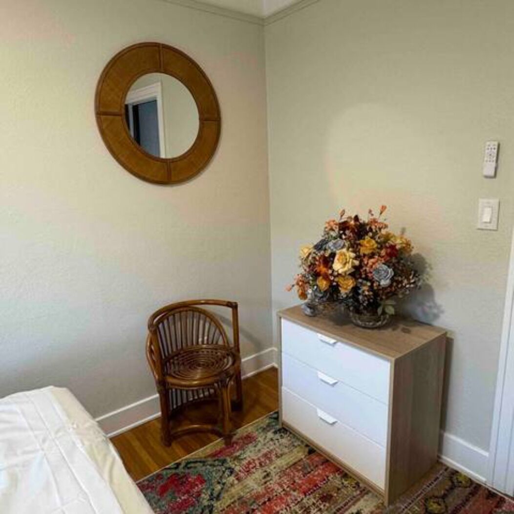A bedroom with a bed, a wooden chair, a dresser with a floral arrangement, and a round mirror on the wall.