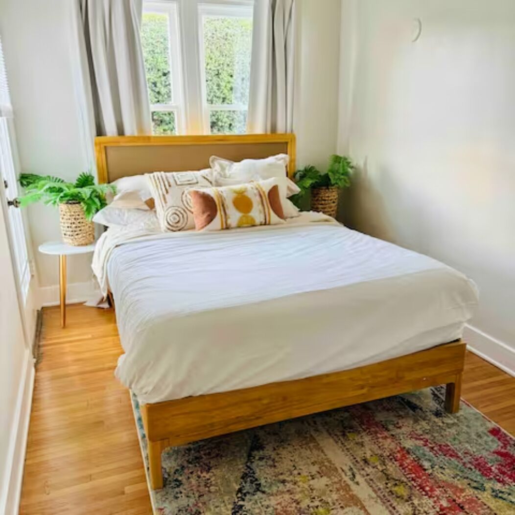 A neatly made bed with a wooden frame, a patterned rug, and two potted plants on either side.