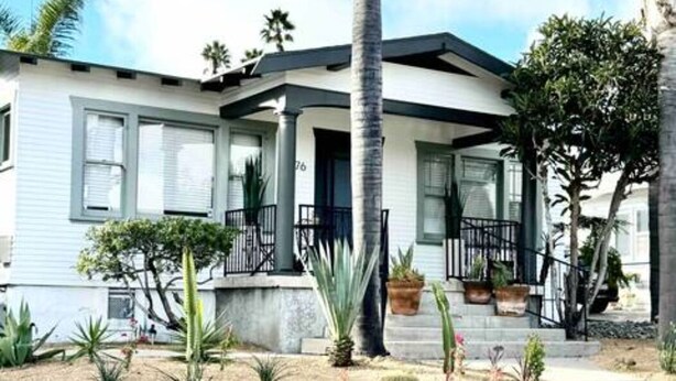 Una casa de dos pisos con un porche delantero, un pequeño jardín con diversas plantas y una palmera.