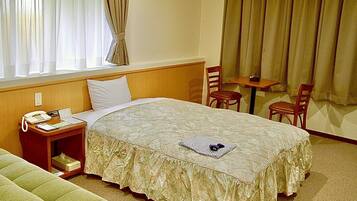 Una habitación de hotel con cama, una mesita de noche con teléfono y lámpara, una pequeña mesa con dos sillas y una ventana con cortinas.