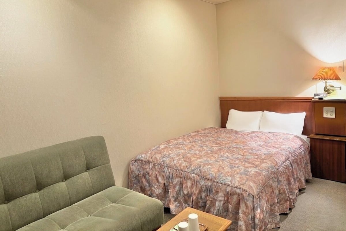 A small, neatly arranged hotel room with a single bed, a green sofa, a bedside table with a lamp, and a wooden nightstand.