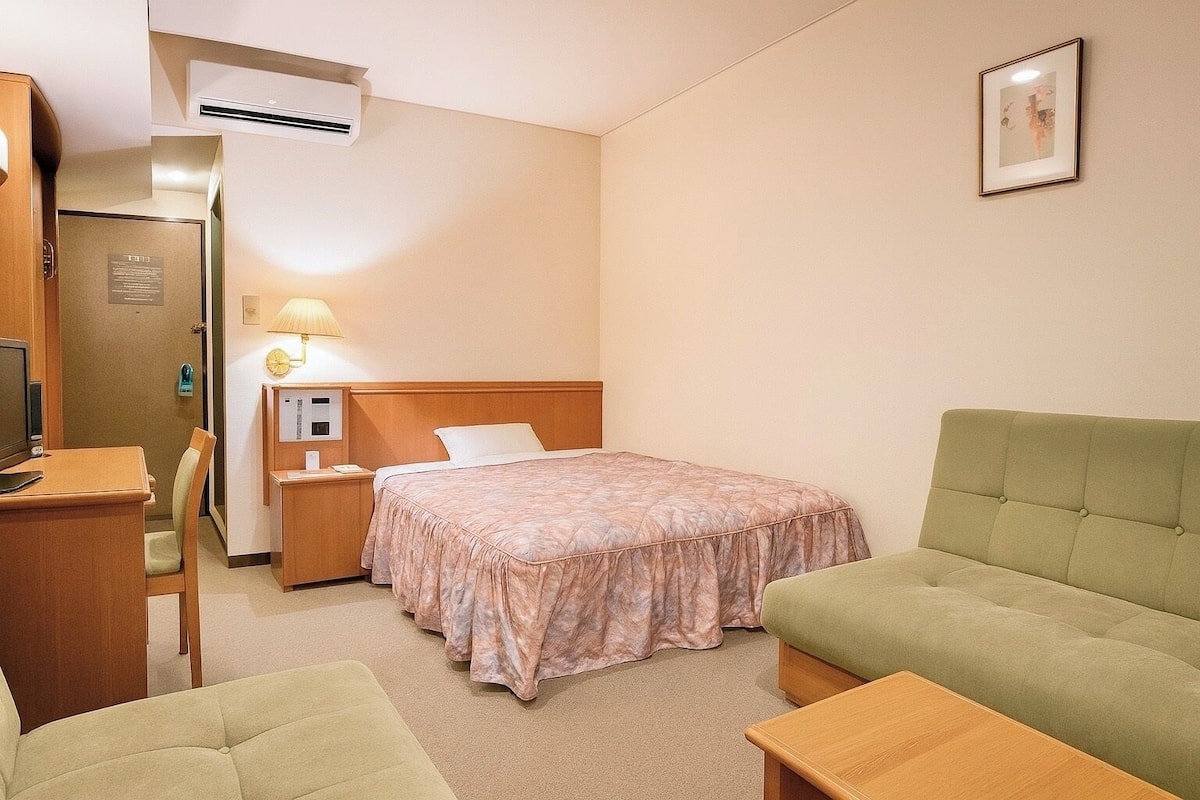 A hotel room with a bed, a desk, a chair, a bedside table with a lamp, and a green sofa.