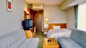A hotel room with two grey sofas, a wooden side table, a television, a lamp, and a bed with a white bedspread. - Matsue Universalhotel   (Matsue)