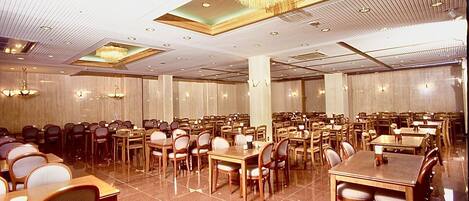 Un amplio comedor de hotel con varias mesas y sillas, grandes candelabros y suelo de baldosas.