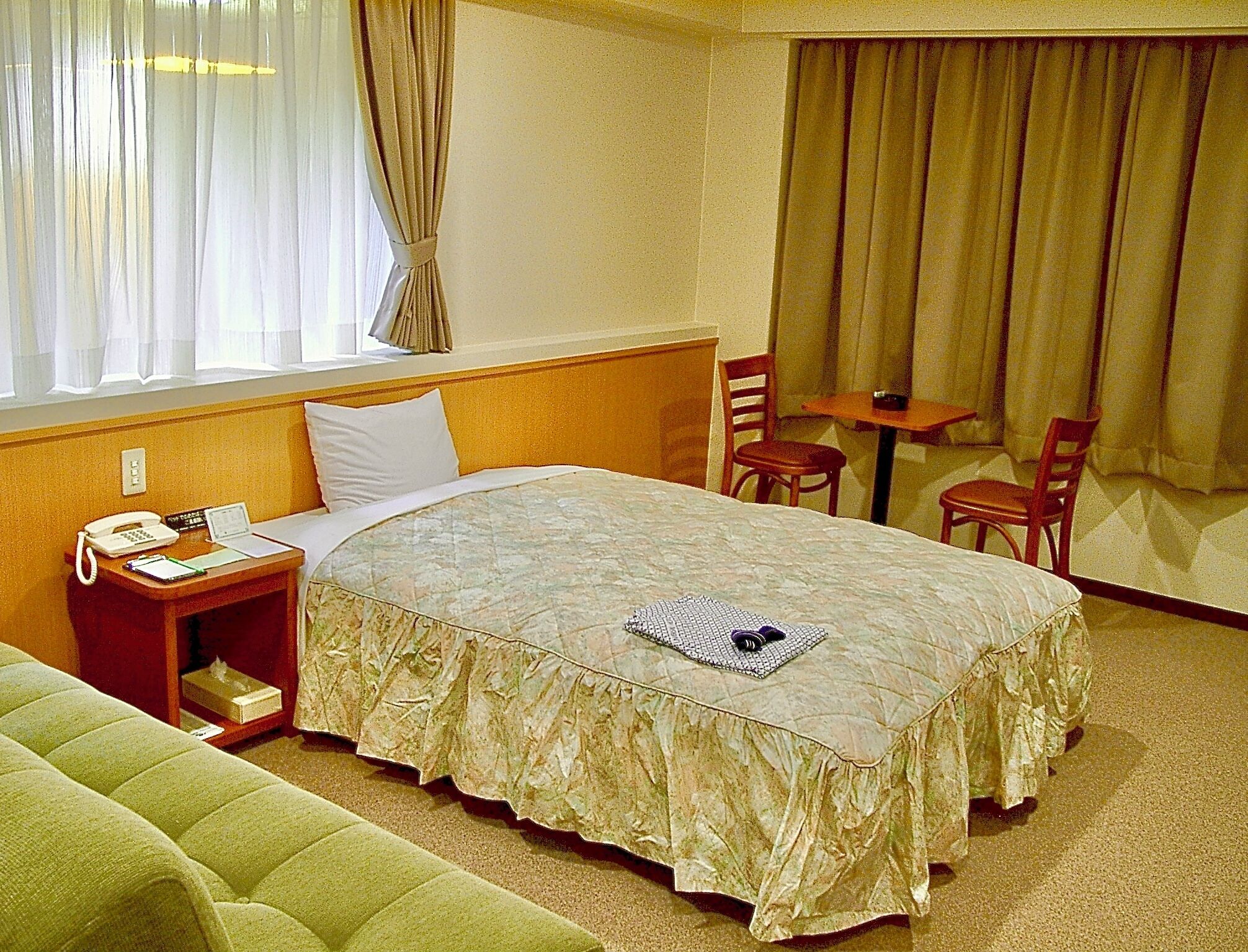 A hotel room with a bed, a bedside table with a phone and a lamp, a small table with two chairs, and a window with curtains.