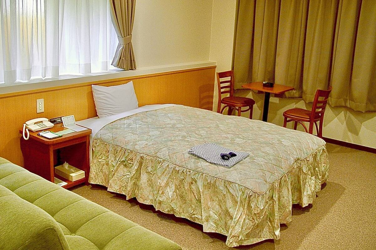 A hotel room with a bed, a bedside table with a phone and a lamp, a small table with two chairs, and a window with curtains.