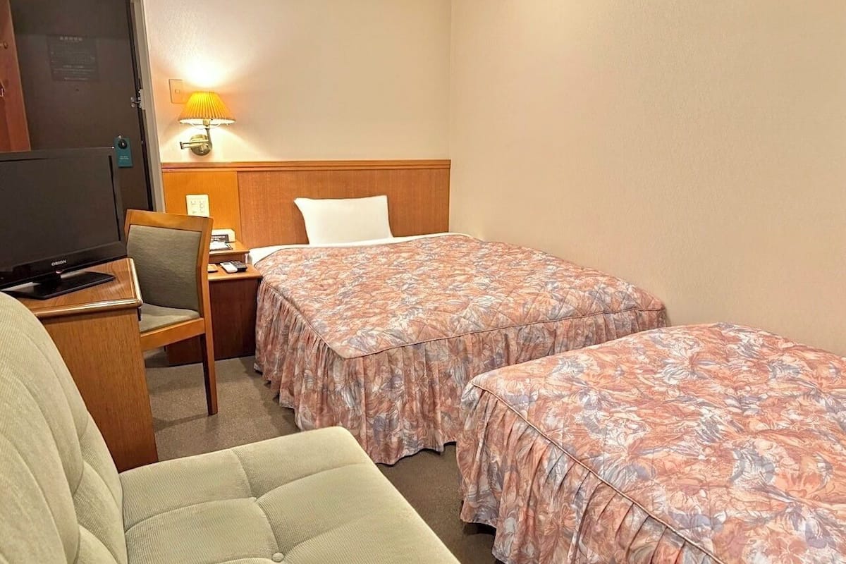 A hotel room with two beds, a desk, a chair, a television, and a lamp.