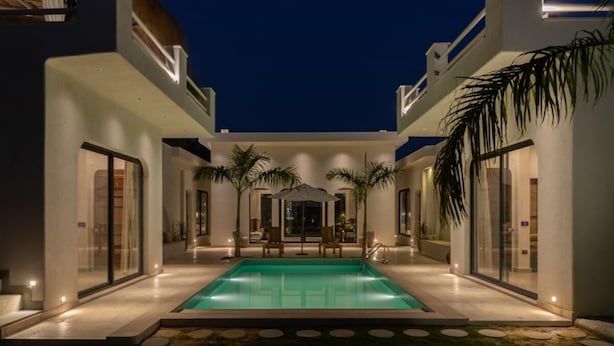 Uma área de piscina moderna com piscina retangular, rodeada por portas de vidro e palmeiras, sob um céu escuro.