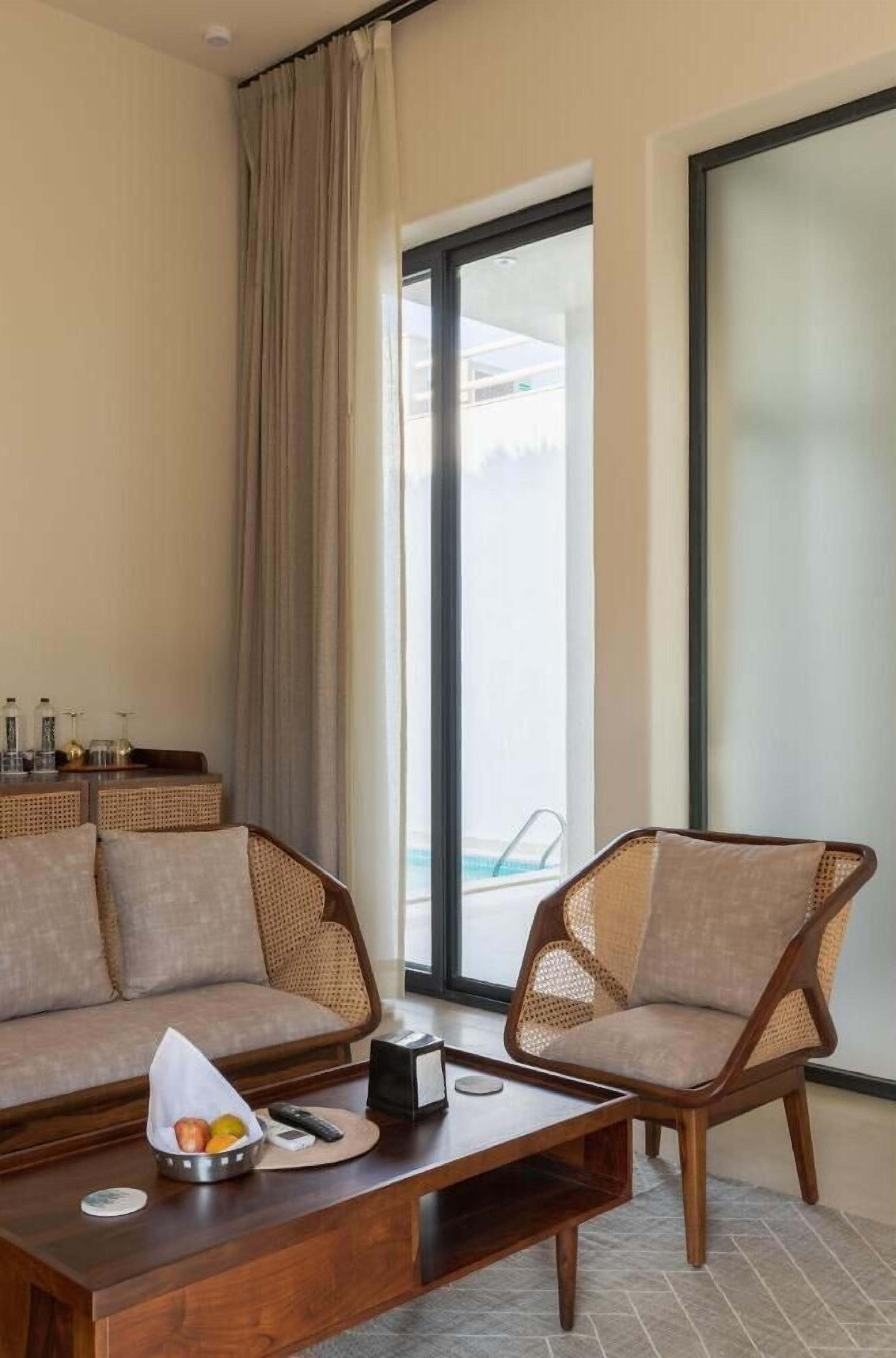 Habitación de hotel con sofá, sillón, mesa de centro con frutero y vista a una piscina a través de un ventanal grande.