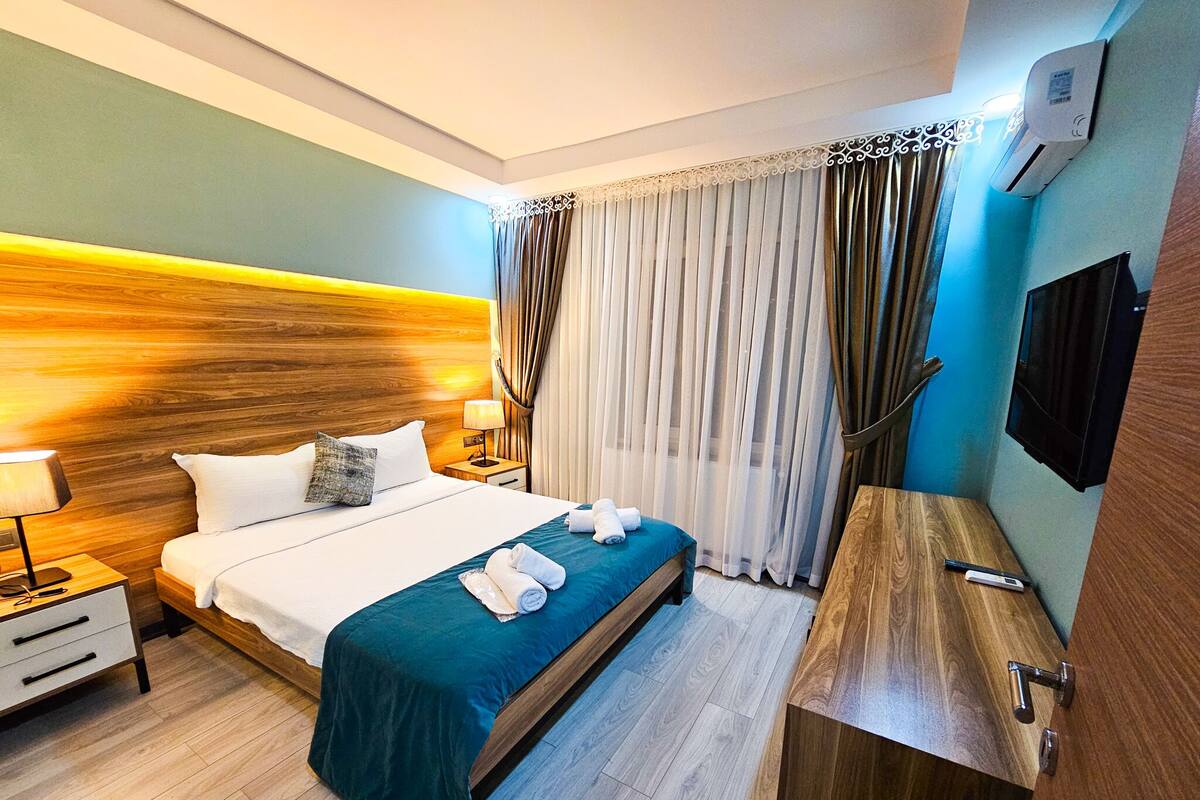 A hotel room with a wooden headboard, a bed with a teal bedspread, a bedside table with a lamp, a wooden desk, and a TV mounted on the wall.